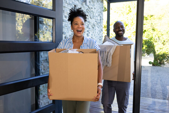 African American couple smiling while carrying moving boxes with blanket and cushion into home