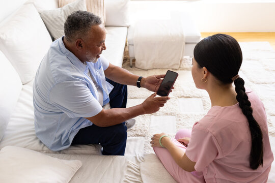Diverse senior man and woman sitting on white sofa and rug, holding smartphone and pointing