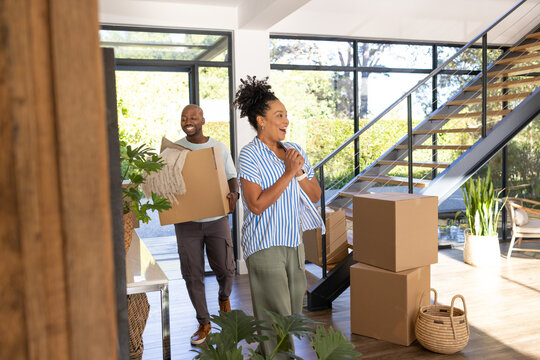 Couple moving into modern home entry carrying cardboard box with folded blanket near staircase