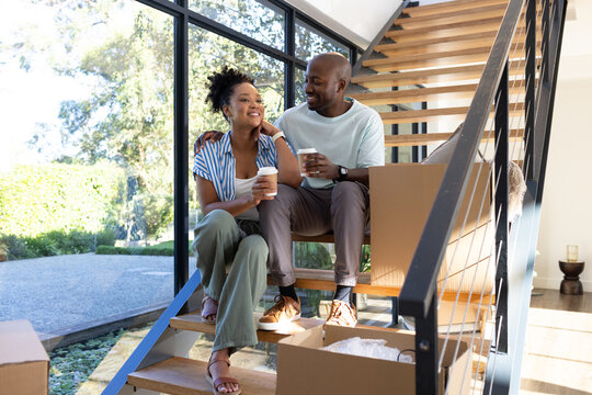 African American couple sitting on staircase at modern home amid moving boxes holding coffee cups