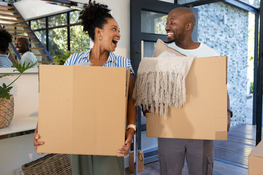 African American couple carrying cardboard moving boxes with blanket, pillow smiling in home foyer
