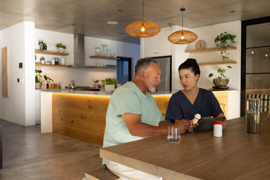Asian nurse in scrubs showing pill bottle, tapping tablet beside patient at kitchen table