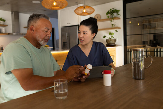 Senior African-American man and Asian RN in scrubs checking meds at island using tablet pill bottle