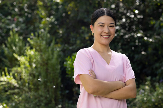 Asian woman standing with arms crossed, wearing pink medical scrubs and hoop earrings in garden