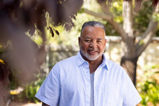 Senior African American man standing in sunlit backyard wearing light blue shirt, framed by leaves