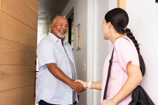 Senior African American man and Asian carer greeting at front door shaking hand wearing pink scrubs
