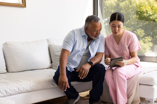 Senior African American man watching Asian nurse in pink scrubs pointing at tablet in living room