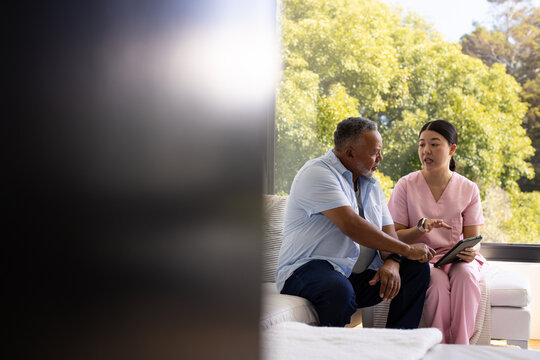 Diverse provider in pink scrubs tapping tablet while mature man with stethoscope listening at home