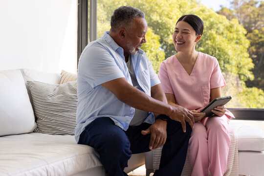 Asian woman nurse in pink scrubs and African American senior sitting on white sofa reviewing tablet