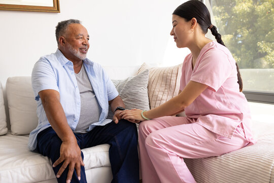 Diverse senior man and female caregiver holding hands on sofa in pink scrubs at home