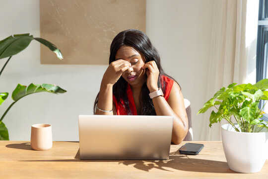African woman holding phone to ear and pinching bridge of nose at home office with laptop