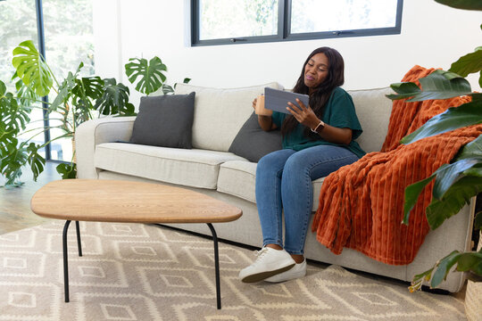African American woman sitting right sofa at home in green-top holding tablet and mug, copy space