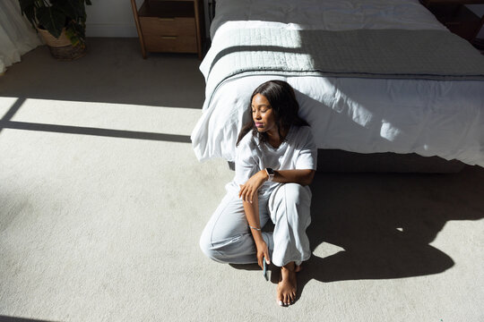 African woman sitting beside bed in bedroom wearing gray pajamas holding smartphone, copy space
