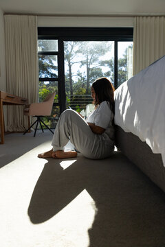 African woman sitting on floor in bedroom, leaning against bed, holding smartphone by window