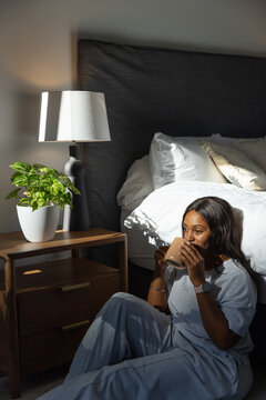 African woman sitting beside bed sipping tan mug in grey set, lamp, plant, sunlight casting shadows
