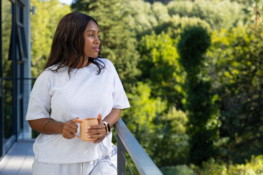 African woman wearing light gray sweats standing on balcony holding beige mug, gazing toward trees