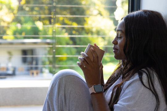 African American woman sitting by window holding mug wearing loungewear and smartwatch, copy space