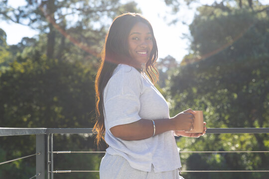 Adult African woman standing on balcony by metal rail holding ceramic mug, wearing gray loungewear