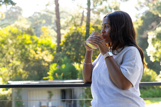 African American woman leaning on balcony railing sipping from glass wearing smartwatch, copy space