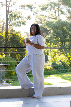 African American woman standing barefoot on terrace holding mug wearing gray loungewear, smartwatch