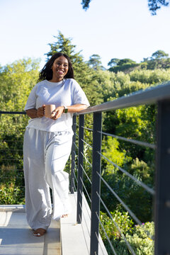 Woman standing barefoot leaning on metal balcony railing, holding ceramic mug, wearing grey sweats