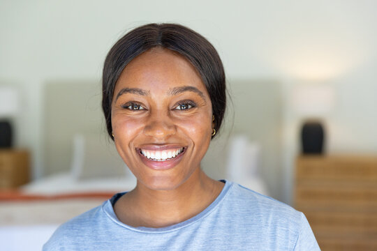 Female smiling, sitting in bedroom wearing blue shirt with bed and bedside lamp visible