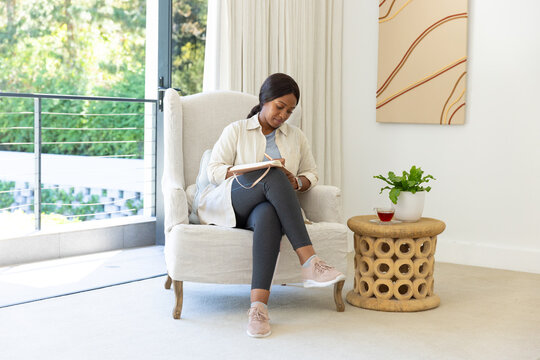 African American woman sitting in armchair writing in notebook with pen by balcony in pink sneakers