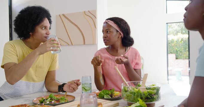 African American friends sitting at counter, starting chat; third reaching for pizza causing smiles