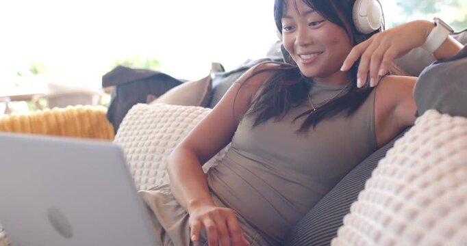 Asian adult woman reclining on couch reacting to laptop content while adjusting headset and smiling