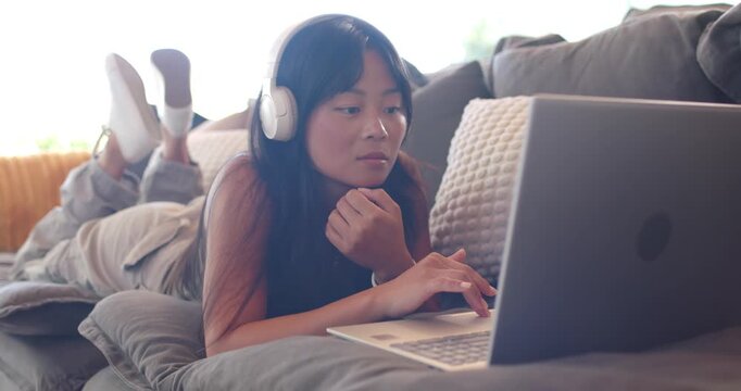 Asian woman lying on couch using laptop with headphones, laughing after receiving funny text
