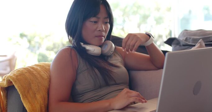 Asian woman working on laptop, adjusting headphones then checking smartwatch on couch near window