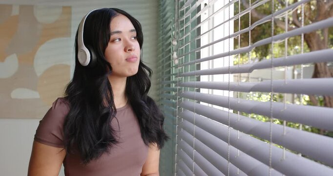 African American woman listening on white headphones, standing near blinds, glancing at trees