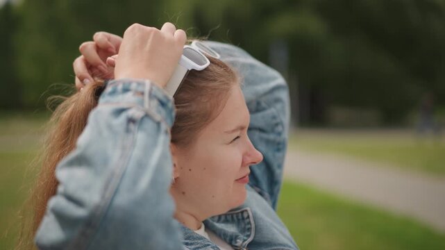 Caucasian student tying ponytail outdoors denim jacket, white sunglasses perched on head, hands gathering hair, pathway and greenery in background, soft sunlight, thoughtful expression
