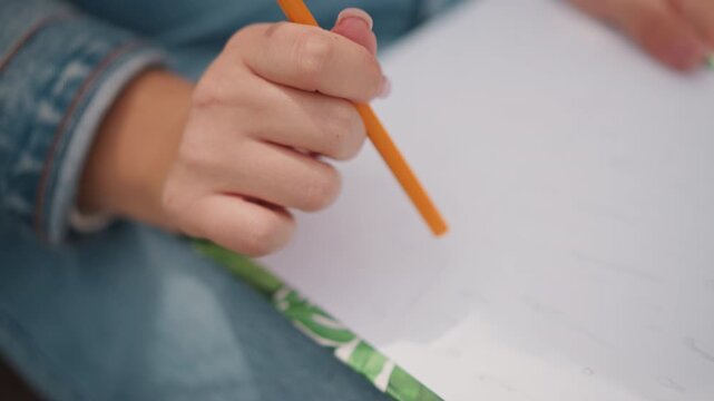 Child hand holding pencil on paper intimate close up of young student wearing denim jacket gripping orange pencil above patterned notebook careful handwriting practice during quiet homework session