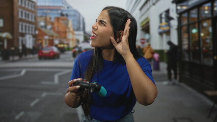 Young hispanic woman holding a gamepad cups her ear while listening and looking puzzled amid busy storefronts and passing cars on a street  curiosity focus. © Krakenimages.com