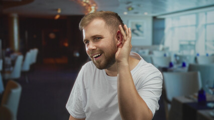 Obraz premium Man cupping hand to ear listening in building dining area wearing white t shirt and beard; curiosity listening engagement.