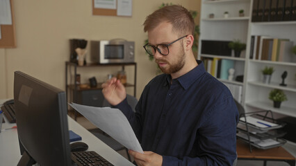 Fototapeta premium Man reading printed document and holding paper at a building office desk near computer monitor and shelves; concentration paperwork.