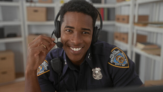 Man police officer smiles and adjusts headset microphone while speaking at a desk in a building with shelves and boxes, badge visible; professional duty readiness.