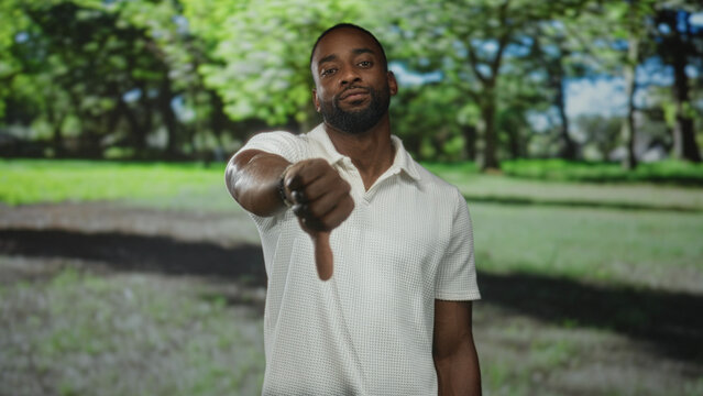 Young african american man extending thumb down toward camera in sunlit forest park with blurred trees behind; disapproval.