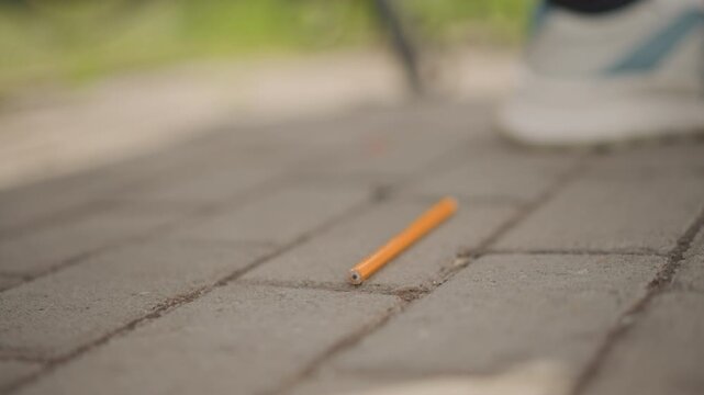 Small object lands on pavement quietly. Brief slip causes pencil to land unexpectedly on path. An orange pencil accidentally drops onto brick surface near sneaker in tranquil park