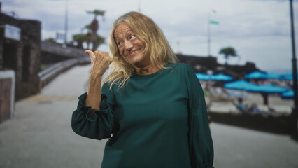 Obraz premium Senior woman points thumb to street beside beach umbrellas while smiling and showing hand; contentment.