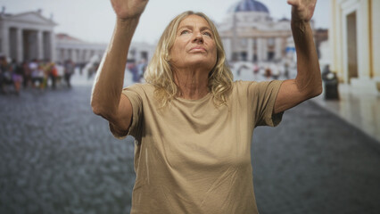 Senior blonde woman raising both arms with bare forearms visible in a busy piazza before a basilica, looking upward  quiet contemplation. © Krakenimages.com