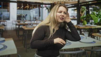 Fototapeta premium Woman holds hand to ear, wincing with closed eyes while leaning toward set table and plates in a busy restaurant building; frustration.
