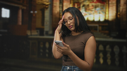 Obraz premium Woman holding smartphone and scrolling with fingers while looking down in a building with ornate stained glass; concern contemplation.