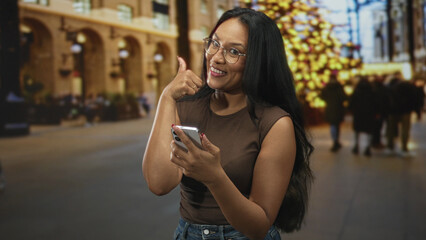 Fototapeta premium Woman tapping smartphone screen and giving a thumbs up on a city street by a decorated building and large christmas tree; holiday joy.