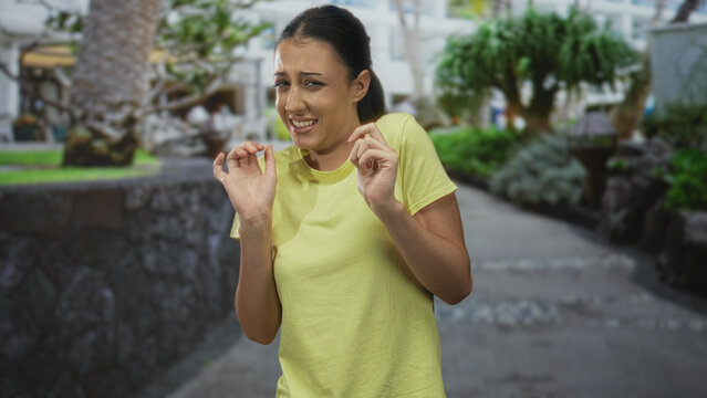 Young hispanic woman with hands raised recoiling on a street walkway wearing a yellow t shirt; disgust aversion.