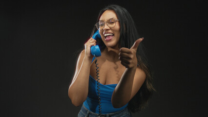 Obraz premium Woman talking on a vintage blue corded telephone, hand to ear and smiling broadly while reaching forward in studio; joyful connection.