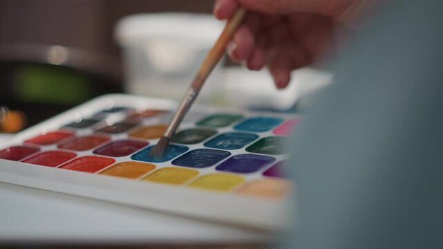 Closeup watercolor palette brush dipping into vibrant pans, studio tabletop with ceramic cup and jars, focused illustrator selects teal pigment, soft artificial lighting, precise strokes