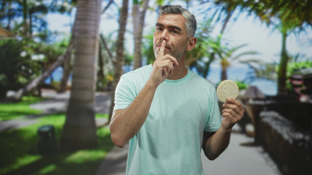 Man with grey hair holds a round corn pastry and shushes with finger to lips on a seaside street lined with palm trees; playful secrecy.