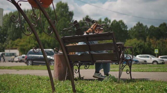 Wide shot bench roadside caucasian commuter waiting, traffic and parking lot backdrop, planter and ironwork frame, cloudy sky over treeline, jeans and sneakers visible, midday observational pause